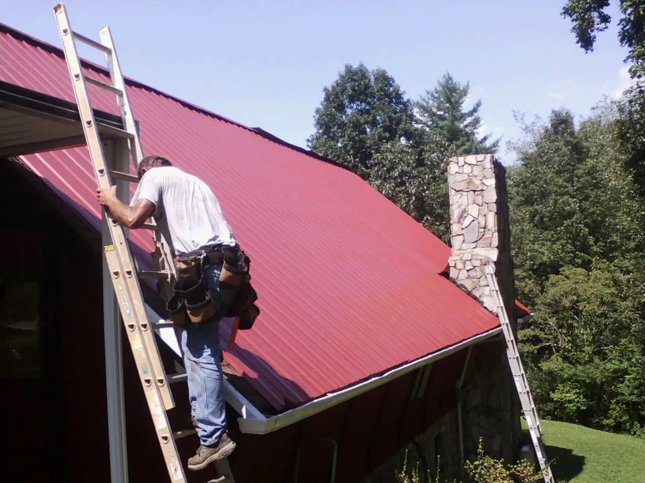Roofer on a ladder beside a red metal roof and stone chimney — Soffit & Fascia Repair by Roofing Brothers in Allen